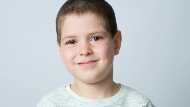 Ένα χαριτωμένο 5χρονο αγόρι χαμογελάει, ανοιγοκλείνει τα μάτια, κοιτάζει μακριά σε ένα λευκό φόντο - Πλάνα, βίντεο