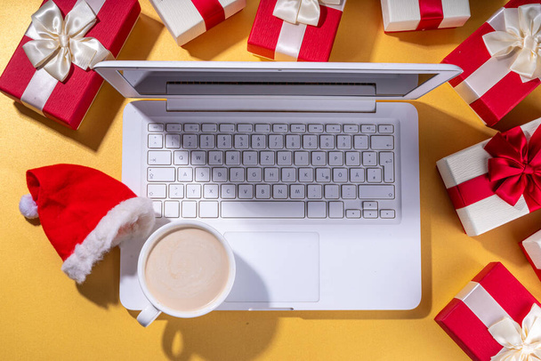 Noel indirimi, tatil hazırlıkları. Noel Baba 'nın elleri dizüstü bilgisayarlı, sıcak çikolatalı latte bardağı, hediye kutuları, altın arka planda yatıyordu. Dilek listesi yapıyorum, Noel için hazırlık hediyeleri hazırlıyorum.. - Fotoğraf, Görsel