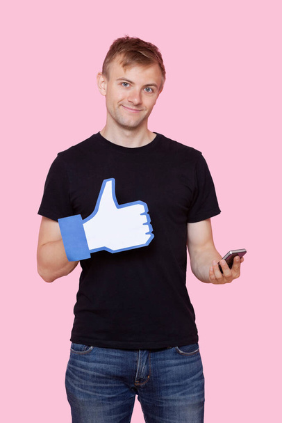 "Portrait of a happy young man with cell phone holding fake like button over pink background" - Photo, Image
