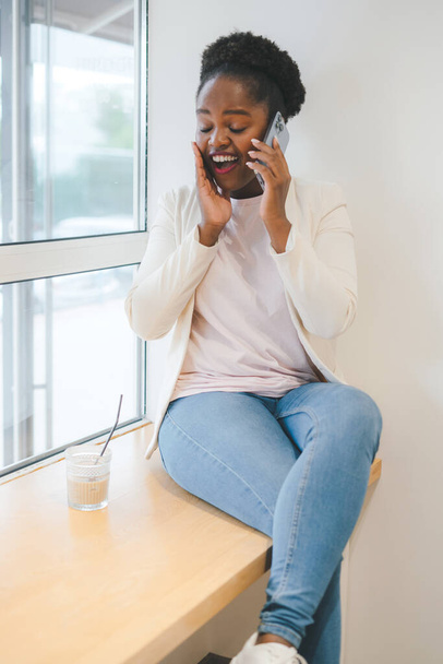 Gülümseyen Afrikalı genç bir kadın kafede oturmuş telefonda konuşuyor ve iyi haberler alıyor. Kapalı bir kafede dinlenmek, cep telefonu kullanmak, konuşmak. - Fotoğraf, Görsel