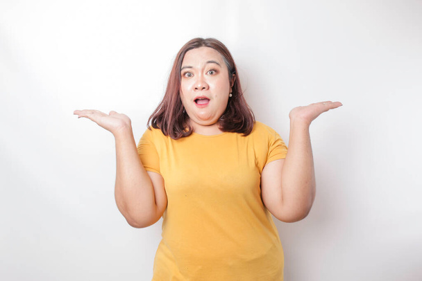 A portrait of an Asian big size woman wearing a yellow shirt shrugging her shoulder looks so confused, isolated by a white background - Foto, immagini