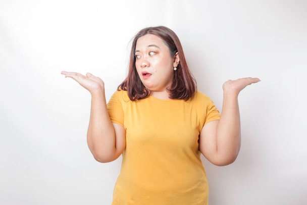 A portrait of an Asian big size woman wearing a yellow shirt shrugging her shoulder looks so confused, isolated by a white background - Zdjęcie, obraz