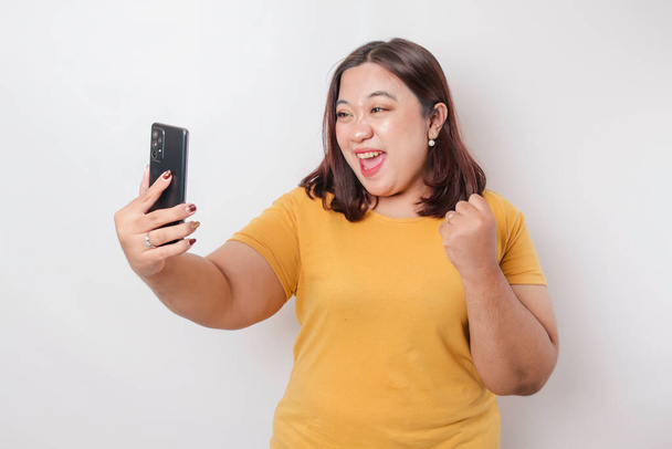 A portrait of a happy Asian big sized woman is smiling and holding her smartphone wearing a yellow t-shirt isolated by a white background - Фото, зображення