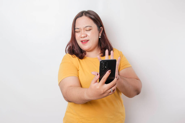 The angry and mad face of big sized Asian woman in yellow shirt while holding her phone on isolated white background. - Foto, Bild