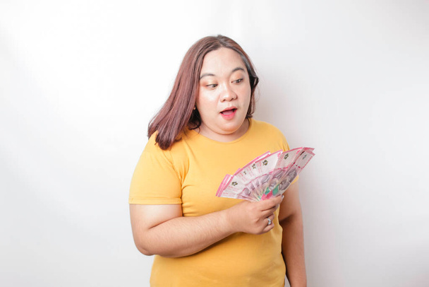 A happy young big size woman is wearing yellow t-shirt and holding cash money in Indonesian rupiah isolated by white background - Foto, imagen
