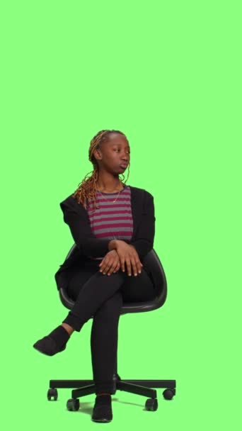 Vertikales Video: Lächelnde Person sitzt auf einem Stuhl in einem Studio mit grünem Hintergrund, wartet und bereitet sich vor. Ungeduldige, fröhliche Frau sitzt auf Hockern und wartet auf die Vorbereitung, Ganzkörper-Greenscreen - Filmmaterial, Video