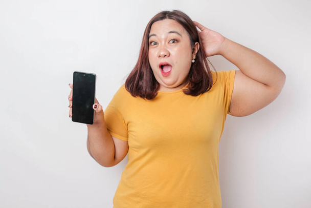 Surprised Asian big size woman wearing a yellow shirt showing copy space on her smartphone, isolated by a white background - Photo, image