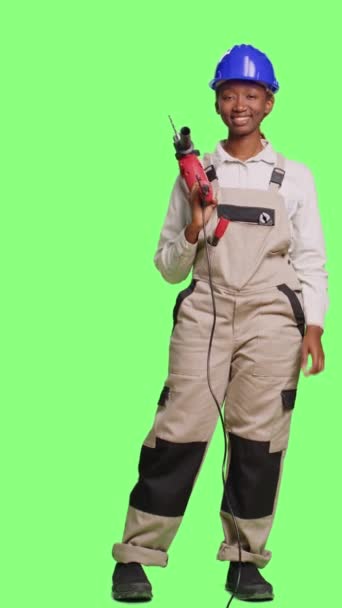 Verticale video: Bouwvakker in overalls en helm met boor- of spijkerschroefpistool op de achtergrond van het volledige groene scherm. Met behulp van boorpistool te renoveren en opknappen, het dragen van uniform - Video