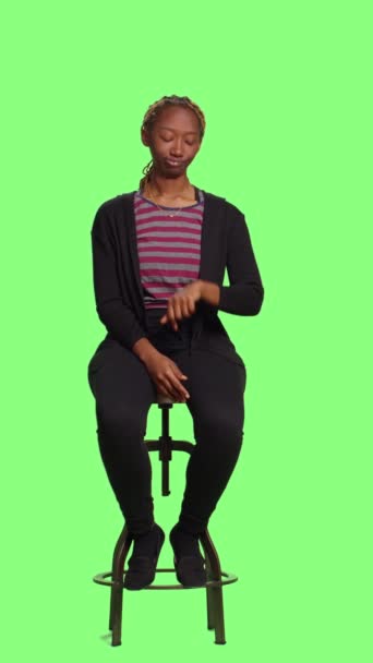 Vertikální video: Nenucená osoba sedící na židli ve studiu nad celotělovou zelenou obrazovkou na pozadí, čekající ve frontě a připravující se. Uvolněná žena sedí v přípravě na zeleném pozadí obrazovky. - Záběry, video