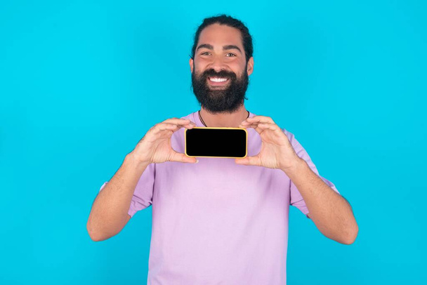 Cheerful cheery content Caucasian man with beard wearing violet T-shirt over blue background holding in hands device hobby smm post blog - Foto, Imagem