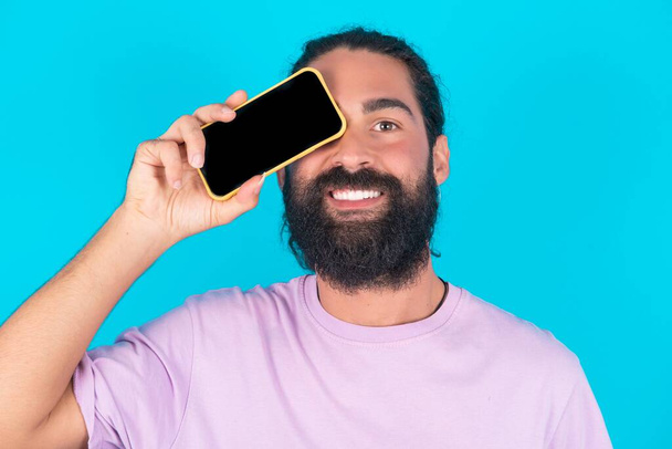 Homme caucasien avec barbe portant T-shirt violet sur fond bleu tenant smartphone moderne couvrant un oeil tout en souriant - Photo, image