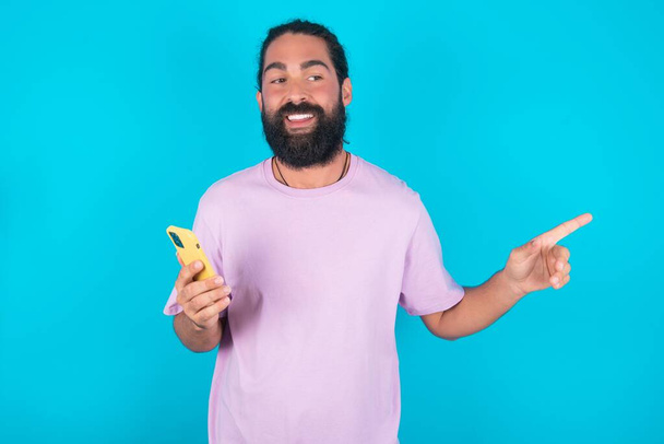 Verblüffter kaukasischer Mann mit Bart trägt violettes T-Shirt über blauem Hintergrund, hält ihr Telefon und zeigt mit dem Finger zur Seite auf leeren Kopierraum - Foto, Bild
