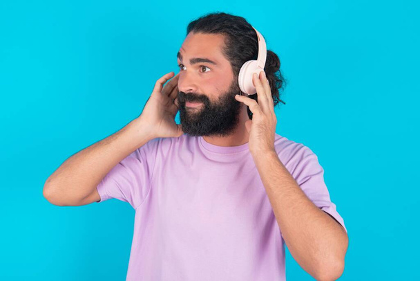 Un homme caucasien barbu portant un T-shirt violet sur fond bleu porte un casque stéréo écoute de la musique concentrée à part. Les gens hobby concept de style de vie - Photo, image