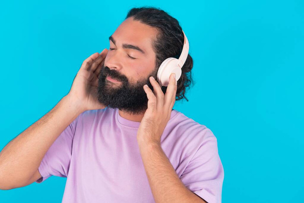 Caucasian man with beard wearing violet T-shirt over blue background  with headphones on head, listens to music, enjoying favourite song with closed eyes, holding hands on headset. - Foto, Imagen