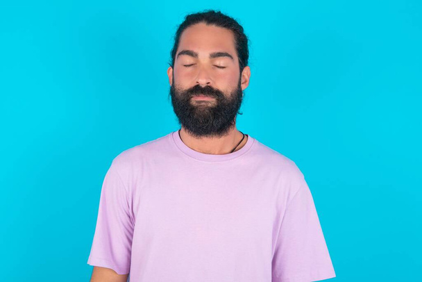 Blanke man met baard draagt violet T-shirt over blauwe achtergrond mooi uitziende zoete charmante schattig mooie mooie winsome zoete vredige gesloten ogen - Foto, afbeelding