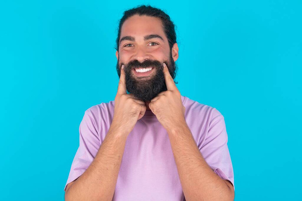 Hombre caucásico feliz con barba vistiendo camiseta violeta sobre fondo azul con sonrisa dentada, mantiene los dedos índice cerca de la boca, los dedos señalando y forzando la sonrisa alegre - Foto, imagen