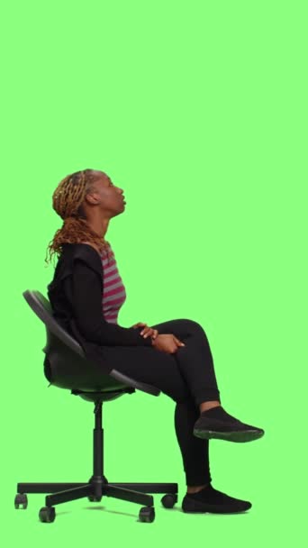 Vertikální video: Mladá osoba na židli sedí a pózuje na zeleném pozadí obrazovky, čeká a připravuje se ve studiu. Netrpělivá žena sedět na stoličce a čekat v přípravě, zelená obrazovka pozadí. - Záběry, video