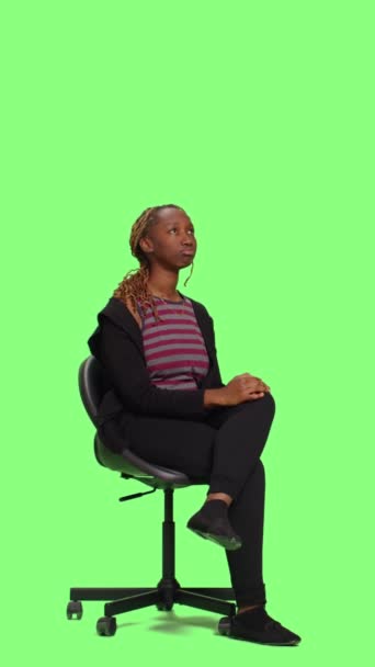 垂直ビデオ:アフリカ系アメリカ人女性がフルボディのグリーンスクリーンを背景に椅子で待って、スタジオで座って準備しています。せっかちな女の子はスツールに座って準備を待って、緑の画面 - 映像、動画