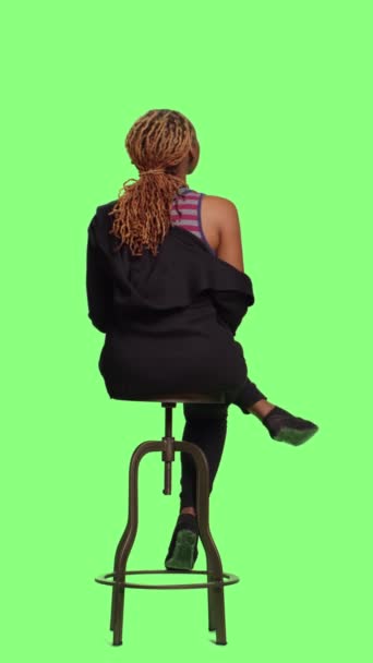 Вертикальне відео: африканська американка на стільці сидить на задньому плані з зеленим екраном. Доросла людина чекає і готується позувати, чекати і сидіти підготовка. - Кадри, відео