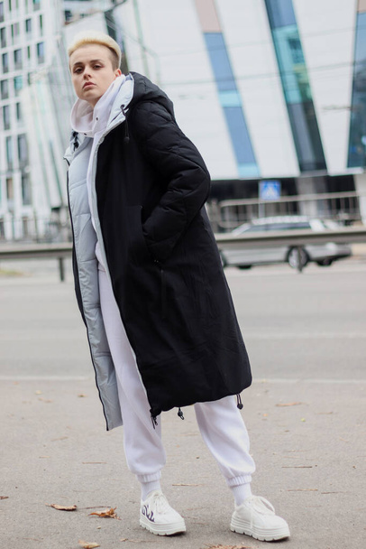 Молодая девушка с короткой стрижкой с светлыми волосами в пальто на фоне высотного здания. Осенняя фотосессия. - Фото, изображение