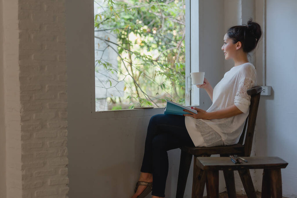 Beautiful young Asian woman drinking coffee and looking through window while sitting on a chair near window at home. - Photo, Image