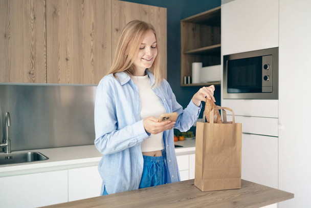 Beautiful young caucasian woman hold eco shopping bag after shopping using smartphone in modern kitchen, delivery service, home purchase in covid-19 lockdown.  - Foto, afbeelding