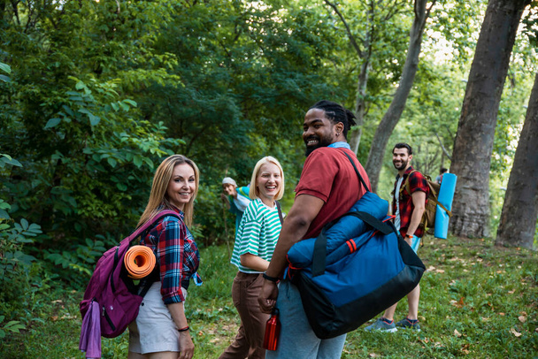 Glückliche erwachsene multiethnische Backpacker, die in Wäldern zwischen grünen Bäumen spazieren gehen. Freunde im Wald. Schmunzelnde Freunde beim Wandern im Wald.  - Foto, Bild