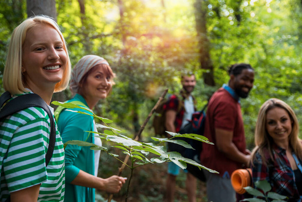 Ευτυχισμένοι ενήλικες Multiethnic backpacker άνθρωποι περπάτημα σε δάση ανάμεσα σε πράσινα δέντρα. Φίλοι στο δάσος. Χαμογελώντας ποικίλες φίλους για πεζοπορία στο δάσος.  - Φωτογραφία, εικόνα