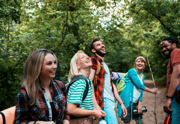 Boldog felnőtt Többnemzetiségű hátizsákos emberek sétálnak az erdőben a zöld fák között. Barátok az erdőben. Mosolygó különböző barátok a túrázás az erdőben.  - Fotó, kép