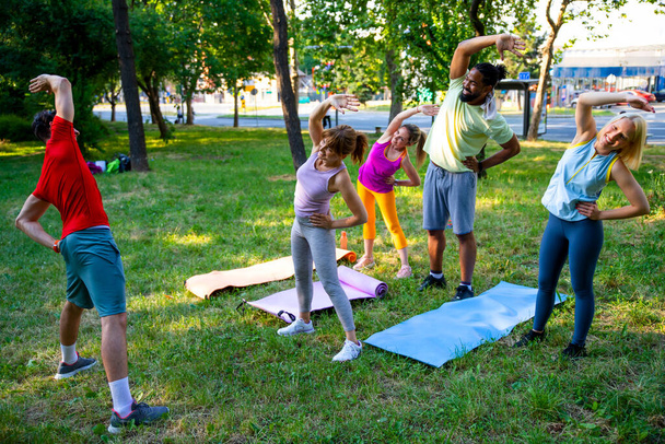 Мультиэтнические друзья занимаются йогой. Люди с тренером занимаются спортом на зеленой траве в парке. Различные люди в фитнес-классе упражнения на открытом воздухе. Тренировка вместе в летний день. Копирование пространства - Фото, изображение