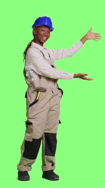 垂直ビデオ:陽気な女性コンストラクターは、広告を行うために背景の上に立って、フルボディの緑の画面上に何かを広告します。背景に例を示す建設労働者は、改装 - 映像、動画