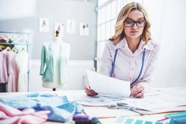 Krásná mladá návrhářka v brýlích drží kousek papíru a zkoumá nějaký náčrt při práci v dressmingu - Fotografie, Obrázek