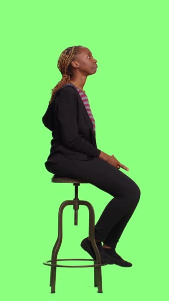 Vertikales Video: Junge selbstbewusste Erwachsene sitzen auf einem Stuhl über einem grünen Bildschirm, warten in der Schlange und bereiten sich vor. Entspannte erwachsene Frauen sitzen in Vorbereitung über Greenscreen-Kulisse im Studio. - Filmmaterial, Video