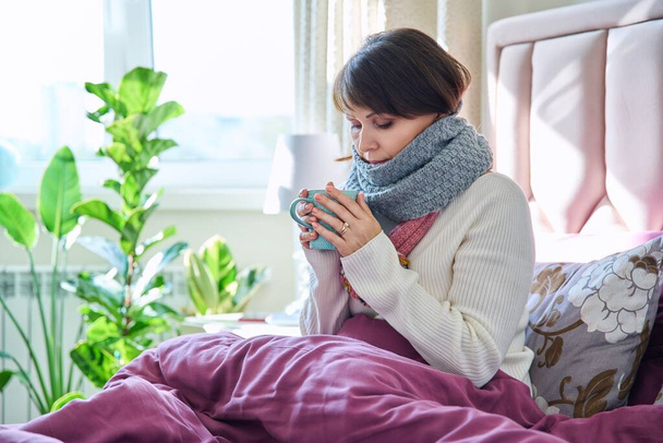Middle aged woman sitting in bed under blanket warming herself with scarf and hot drink in mug, home lifestyle in cold autumn winter season - Photo, image