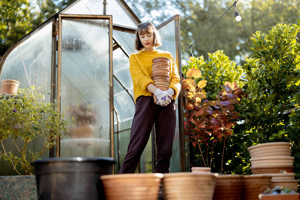 Portrait of a young woman stands with a pile of clay jugs in front of glass orangery at garden in sunny morning. Gardening and hobby concept - Fotoğraf, Görsel