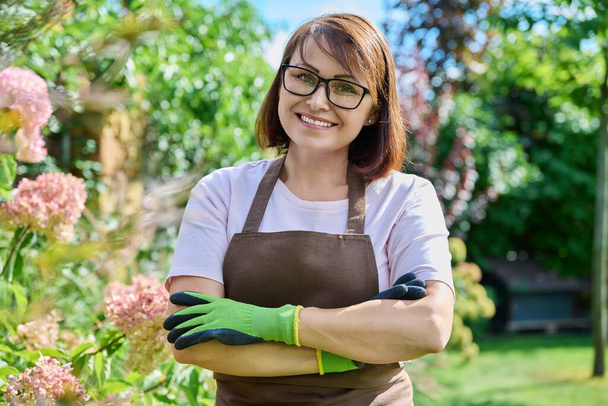 Outdoor portrait smiling female gardener looking at camera in backyard. Confident female in apron wearing gardening gloves with arms crossed, summer garden nature. Work in garden gardening landscaping - Photo, image