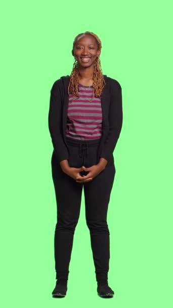 Вертикальное видео: афроамериканец позирует на зеленом экране, стоя всем телом на зеленом экране. Женская модель выглядит естественно и уверенно на изолированном фоне хромы ключа. - Кадры, видео