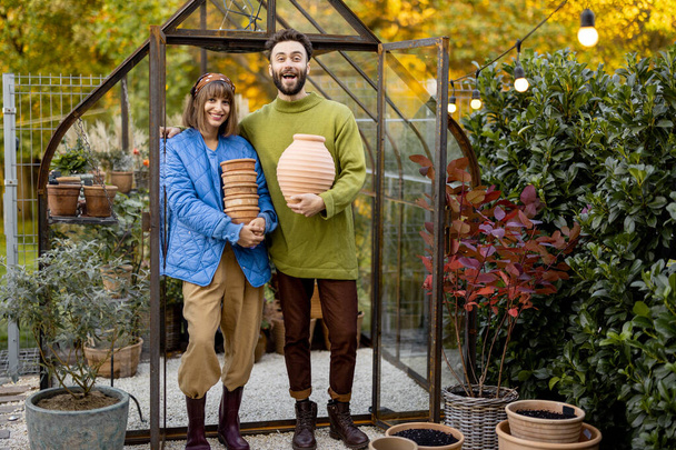 Portrait of a young couple stand together with clay jugs in small greenhouse for growing flowers in garden. Hobby or small family business of growing flowers concept - Photo, image