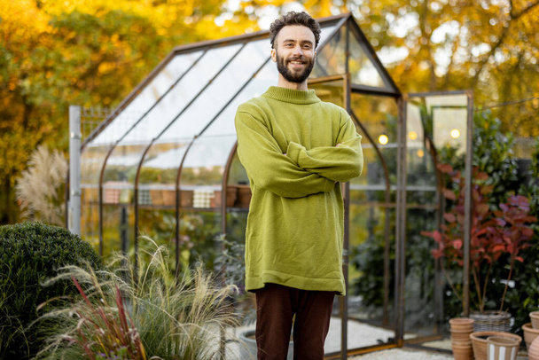 Portrait of a stylish guy stand in front of a tiny greenhouse for growing plants in garden. Hobby, work in garden concept - Foto, Bild