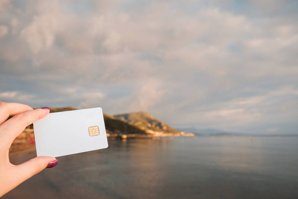 White Bank Card In Woman Hand On Background Of Beach With Sunbeds and Beach Umbrellas In Moraitika, Corfu, Greece. The Concept Of Payment For Relax And Unlimited Possibilities. High quality photo - Foto, imagen