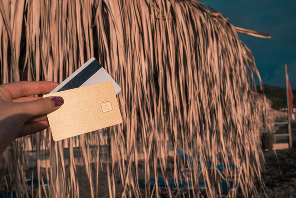 Golden and White Bank Card In Woman Hand On Background Of Beach Umbrellas Made Of Palm Leaves In Moraitika, Corfu, Greece. The Concept Of Payment For Relax, Unlimited Possibilities. High quality photo - Foto, afbeelding