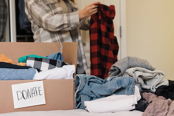 Box with clothing and text donate and woman sorting her clothing. Wardrobe cleaning, donating clothes. - 写真・画像