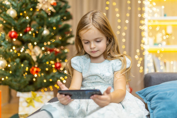 A little girl is watching a video on a smartphone for Christmas, her daughter is celebrating the New Year online near the Christmas tree - 写真・画像