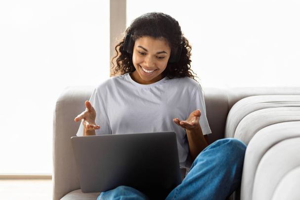 自宅のソファに横たわっている友人とオンラインでウェブカメラに話してノートパソコンを使用してアフロの髪を持つ若いアフリカ系アメリカ人のヒップスター女性。ビデオ通話、バーチャルチャットの概念 - 写真・画像