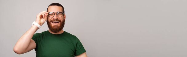 Banner size photo of young handsome man with beard in green t-shirt touching glasses over grey backdrop. - Photo, Image