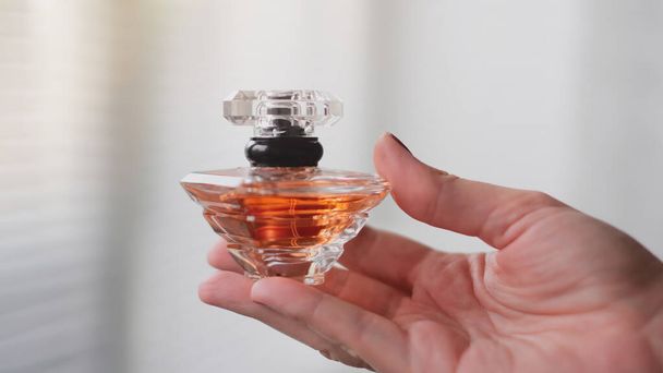 Perfume bottle in a female hand against the background of light room interior. Eau de toilette, eau de parfum, beauty concept. - Photo, Image