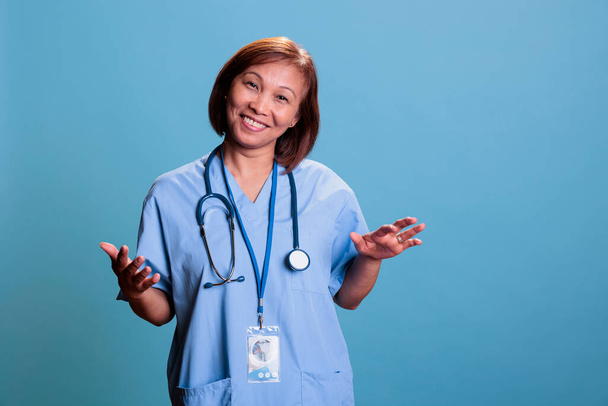 Щаслива азіатська медсестра носить стетоскоп і блакитну форму під час огляду відвідування консультацій, що працюють на лікування. Помічник лікаря, посміхаючись на камеру, що стоїть в студії з синім фоном
 - Фото, зображення