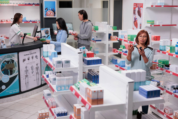 Asian client analyzing boxes and bottles of medicine in pharmacy shop to buy prescription treatment. Looking at pharmaceutical healthcare products, drugstore filled with vitamins and drugs. - Photo, Image