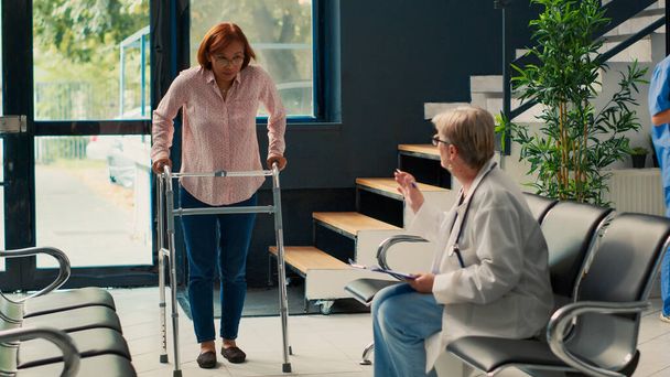 Elderly medic attending physiotherapy with injured patient, doing rehabilitation test and trying to walk with walking frame. Asian woman with leg fracture working on physical recovery. - Photo, Image