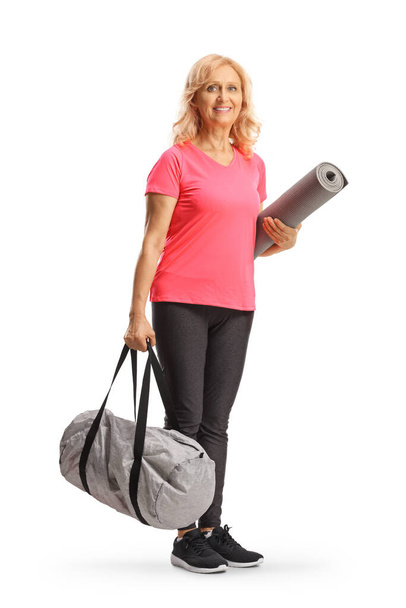 Usmívající se starší žena ve sportovním oblečení drží sportovní tašku a cvičení podložka izolované na bílém pozadí - Fotografie, Obrázek